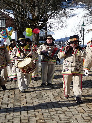 Eine fröhliche Gruppe von Menschen in traditionellen Kleidern, die durch eine schneebedeckte Straße marschiert. Sie spielen Instrumente und tragen bunte Girlanden und Luftballons. | © Hermann Schröttenhamer