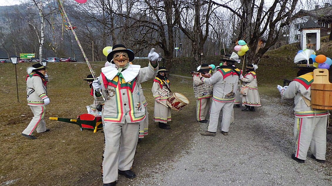 Eine Gruppe von Menschen in traditionellen Kostümen feiert im Freien. Die Teilnehmer tragen bunte Hüte und halten Luftballons, während sie Musik und Freude verbreiten. | © Hermann Schröttenhamer