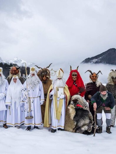 Eine Gruppe von Menschen in traditionellen Kostümen steht im Schnee. Im Hintergrund sind Berge und ein bewölkter Himmel zu sehen. | © Martin Huber