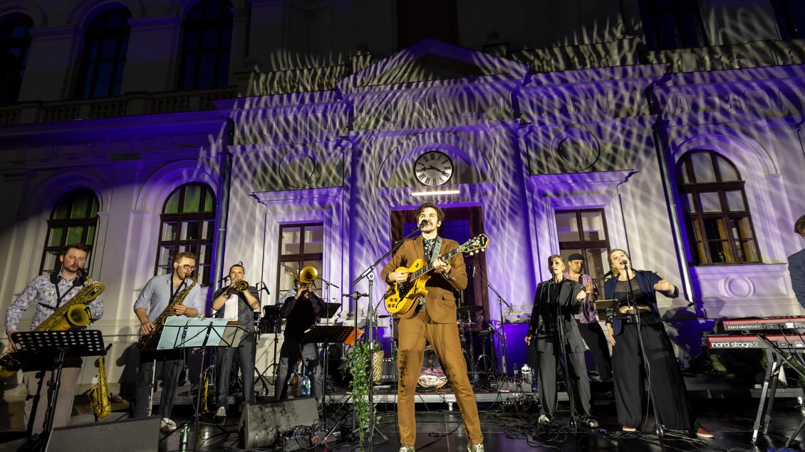 Ein Musiker steht mit einer Gitarre auf der Bühne und wird von einer Band begleitet. Im Hintergrund sind farbige Lichter und eine schöne Architektur zu sehen. | © c_Uni_Graz_Helmut Lunghammer