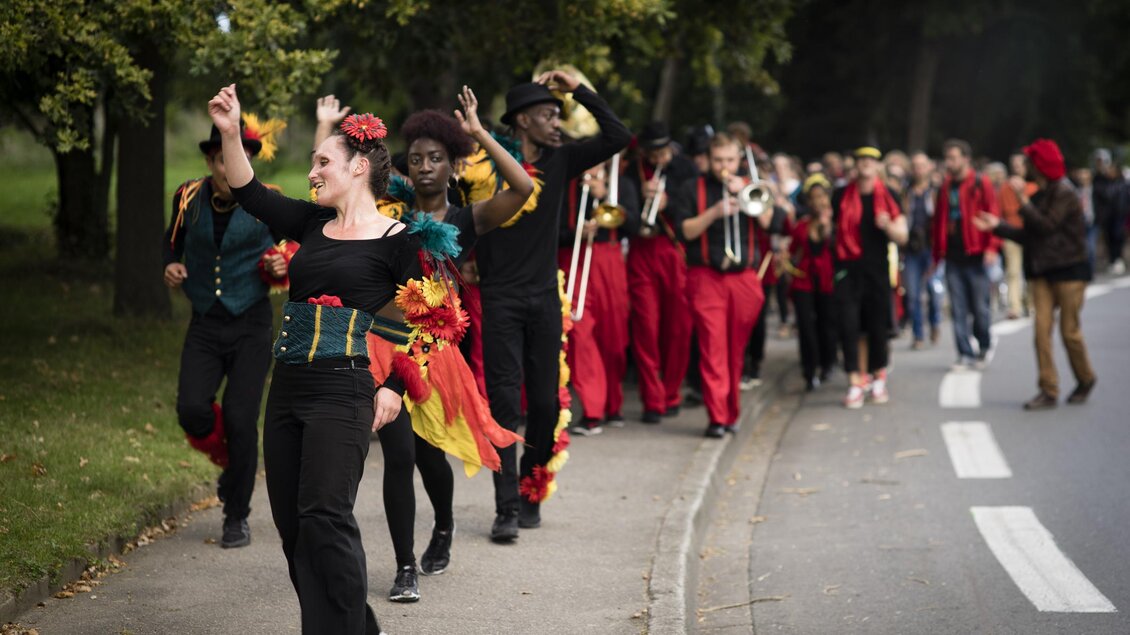 Eine bunte Parade mit fröhlichen Tänzern und Musikern. Die Teilnehmer tragen auffällige Kostüme und ziehen durch eine Straße im Freien. | © _c_Thomas_Guionnet