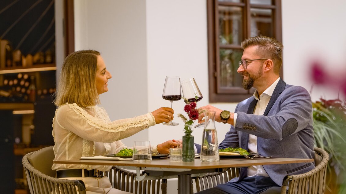Ein Paar genießt ein romantisches Dinner in einem Restaurant. Sie stoßen mit Weingläsern an und haben eine schöne Tischdekoration. | © Der Schrenk