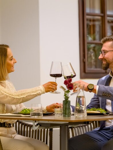 A couple enjoys a romantic dinner in a restaurant. They toast with wine glasses and have a beautiful table decoration. | © Der Schrenk