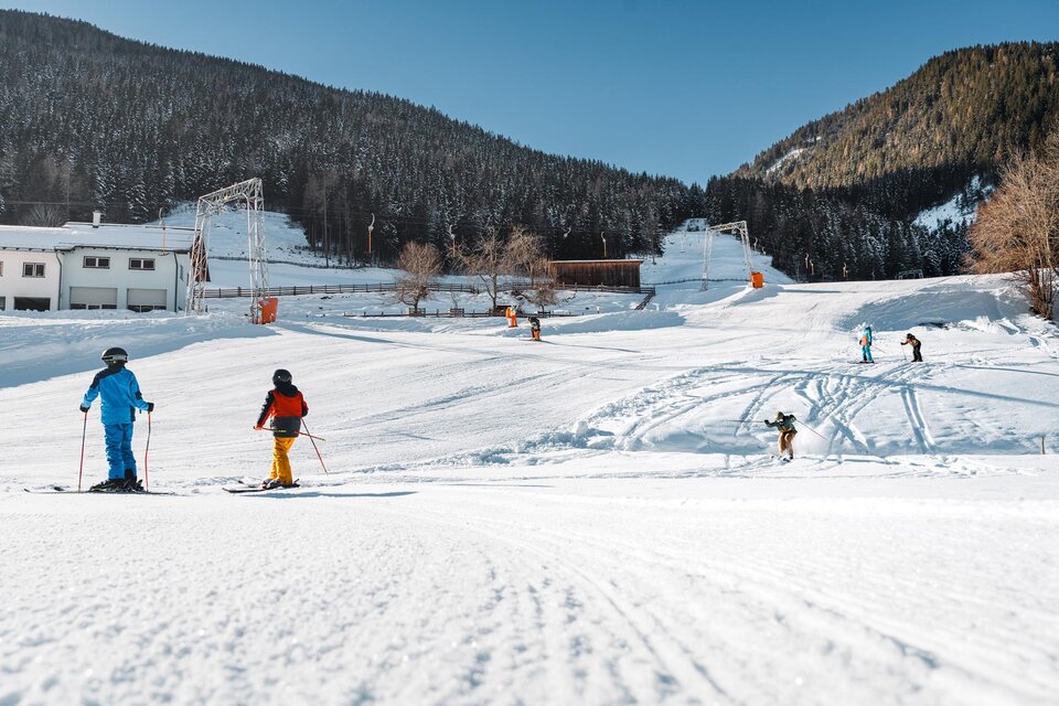 Ein sonniger Skitag in den Bergen mit mehreren Skifahrern auf einer schneebedeckten Piste. Im Hintergrund sind eine Skilift- und Berglandschaft zu sehen. | © Christoph Lukas
