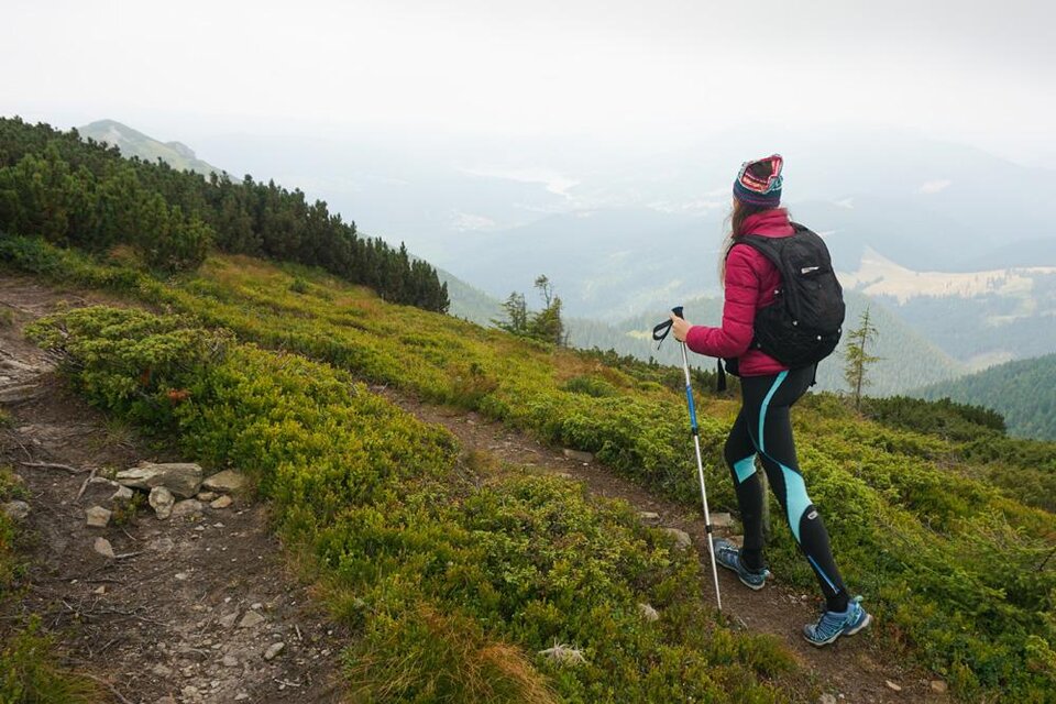 Eine Wandererin steht auf einem Pfad in der Natur. Sie trägt einen rosa Mantel und einen Rucksack, umgeben von grünen Hügeln und einer nebligen Landschaft. | ©  freepik.com