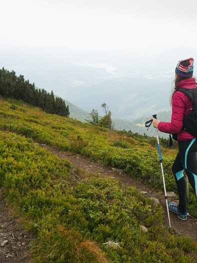 A hiker stands on a path in nature. She is wearing a pink coat and a backpack, surrounded by green hills and a foggy landscape. | ©  freepik.com