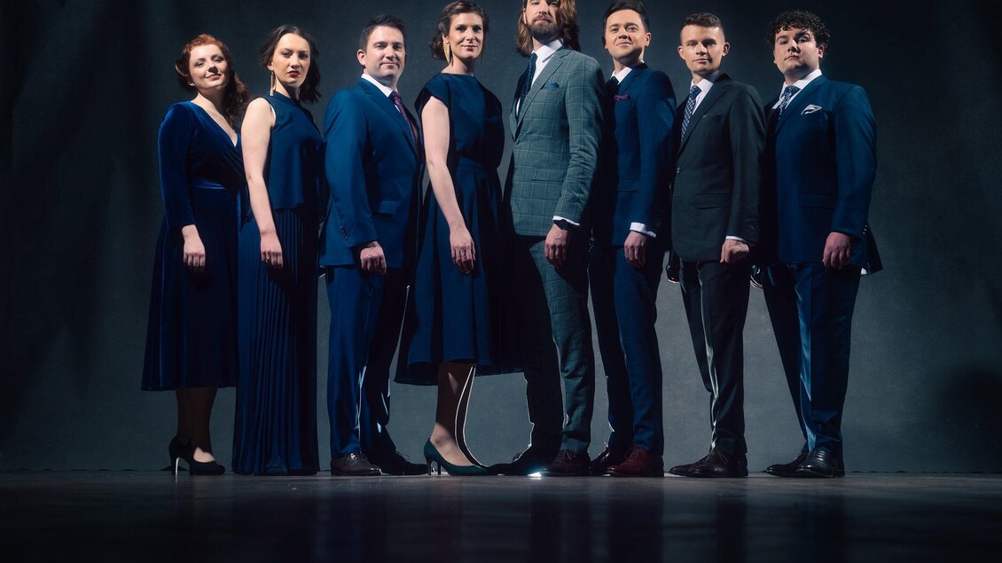 Eine Gruppe von neun Personen steht in eleganter Abendgarderobe auf einer Bühne. Sie lächeln und posieren vor einem dunklen Hintergrund. | © Andy Staples