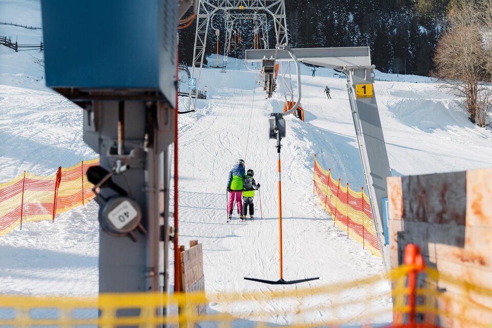 Ein Skilift in einer verschneiten Landschaft. Zwei Personen, ein Erwachsener und ein Kind, bereiten sich darauf vor, den Lift zu benutzen. | © Christoph Lukas