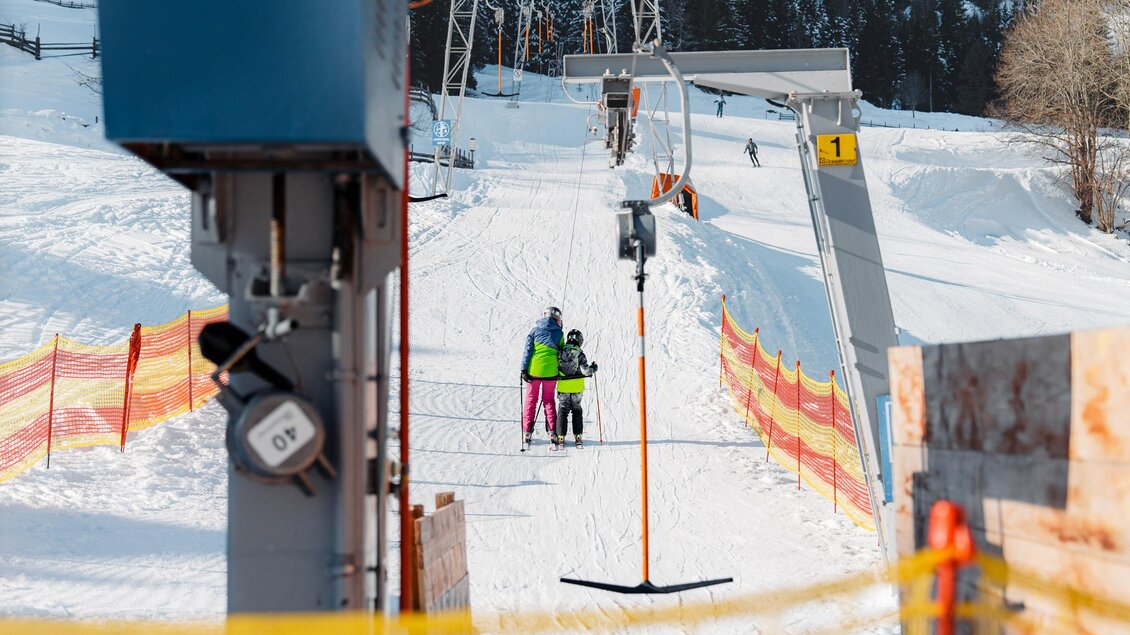 Ein Skilift in einer verschneiten Landschaft. Zwei Personen, ein Erwachsener und ein Kind, bereiten sich darauf vor, den Lift zu benutzen. | © Christoph Lukas