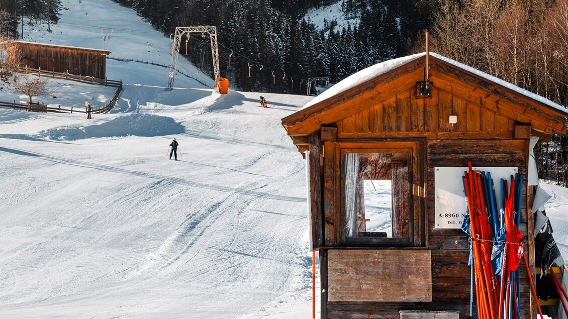 Eine verschneite Berglandschaft mit einem kleinen Holzhaus. Im Hintergrund sind Skifahrer und Lifte zu sehen. | © Christoph Lukas