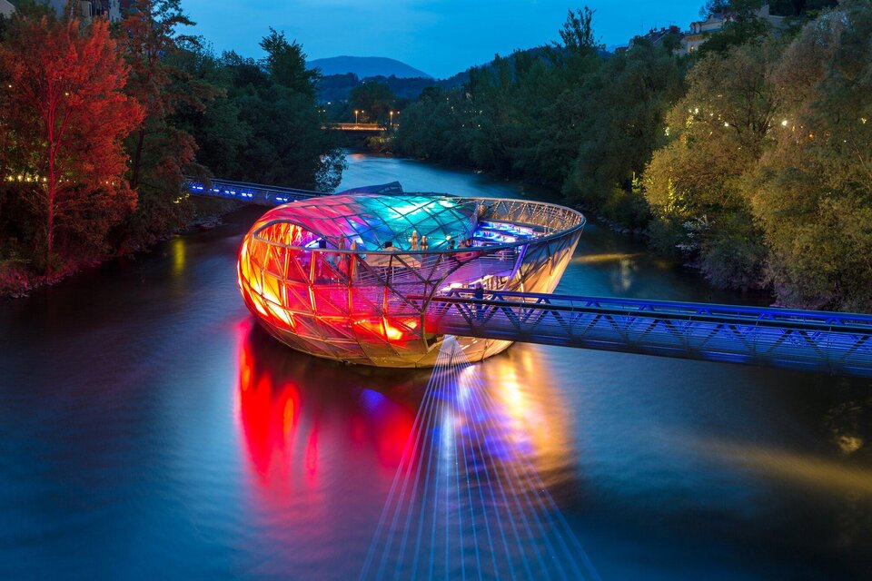 Eine moderne Brücke mit einer ovalen Struktur leuchtet in bunten Farben über einem Fluss. Umgeben von Bäumen und einem klaren Himmel bei Nacht. | © Graz Tourismus - Harry Schiffer