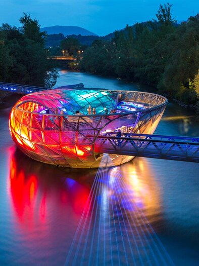 A modern bridge with an oval structure shines in vibrant colors over a river. Surrounded by trees and a clear sky at night. | © Graz Tourismus - Harry Schiffer