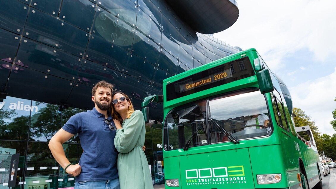 Ein Paar posiert vor einem grünen Bus und einem modernen Gebäude. Die Szene zeigt urbanes Leben und Freizeitaktivitäten. | © Graz Tourismus - Werner Krug