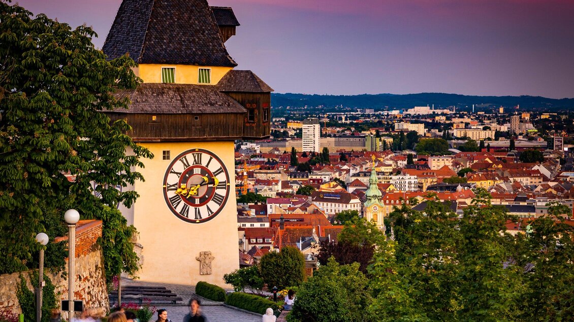 Eine malerische Stadtansicht mit einem alten Uhrturm und vielen bunten Gebäuden. Im Hintergrund erstreckt sich die Landschaft bis zum Horizont. | © Graz Tourismus - Werner Krug