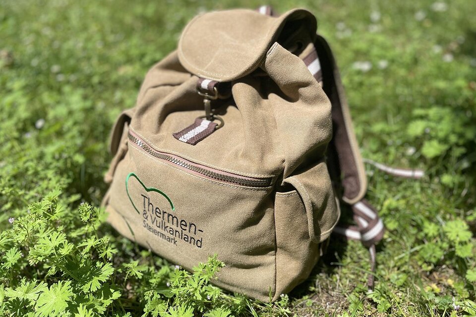 Ein brauner Rucksack liegt auf grünem Gras. Er ist mit einem Logo und einem Reißverschluss ausgestattet. | © TV Thermen- & Vulkanland