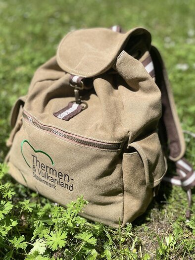 Ein brauner Rucksack liegt auf einer grünen Wiese. Er hat ein Logo von "Thermen-Welt" auf der Vorderseite. | © TVB Thermen- & Vulkanland