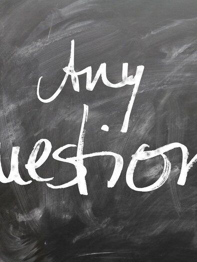 A board with white lettering "Any Questions". The background is dark and slightly messy.