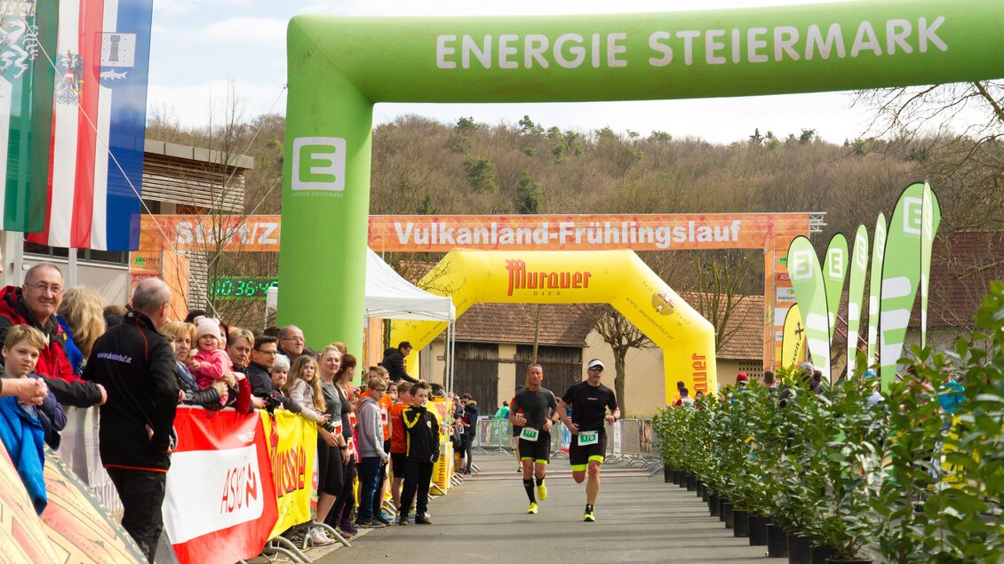Ein Laufereignis mit zwei Läufern, die unter einem großen Bogen mit der Aufschrift „Energie Steiermark“ hindurchlaufen. Zuschauer stehen an der Strecke und feuern die Teilnehmer an. | © Vulkanland Frühlingslauf