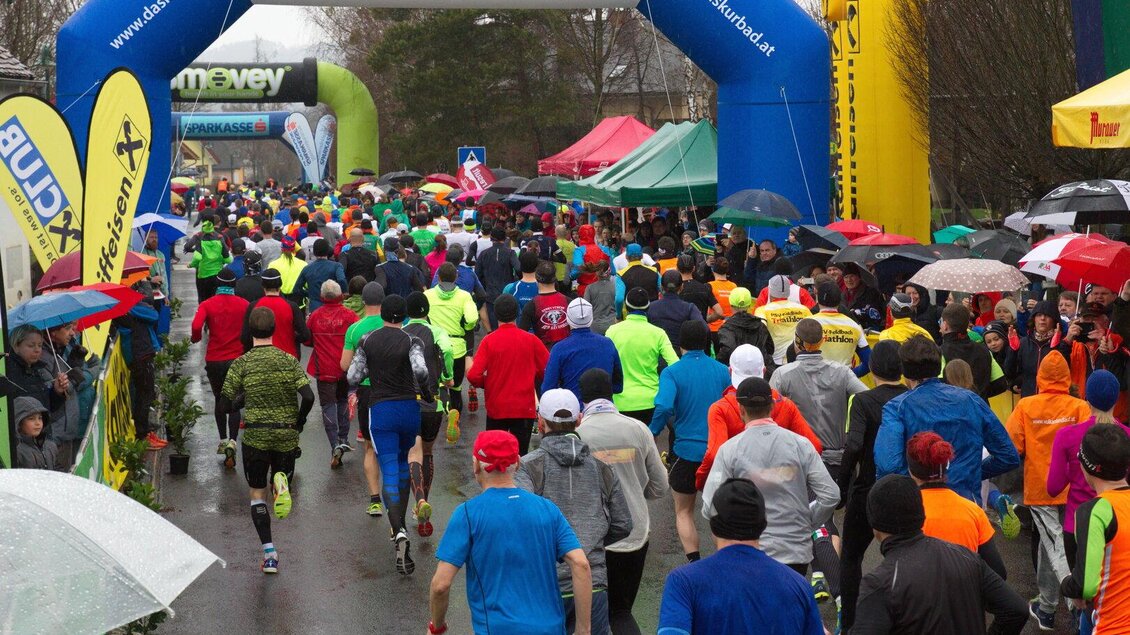 Eine große Gruppe von Läufern in bunten Sportkleidern nimmt an einem Wettlauf bei regnerischem Wetter teil. Im Hintergrund sind Zelte und eine Zielbogen mit der Aufschrift „Kurbad Gleichenberg“ zu sehen. | © ASKÖ Leitersdorf