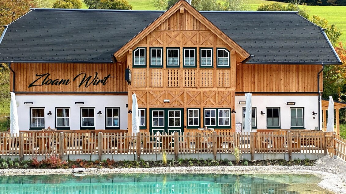 Ein traditionelles Holzgebäude mit einer einladenden Terrasse und großen Fenstern. Im Vordergrund befindet sich ein klarer Teich, umgeben von einer schönen Landschaft.