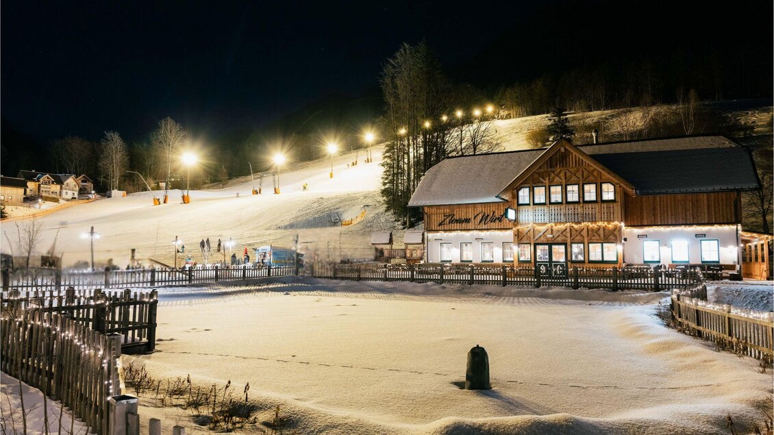 Eine winterliche Landschaft mit einem beleuchteten Skihang und einem rustikalen Holzhaus. Die Umgebung ist mit Schnee bedeckt und es sind Menschen auf den Skipisten zu sehen. | © www.zloam.at