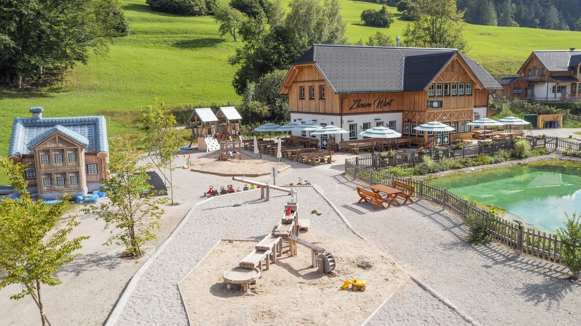 Ein kinderfreundlicher Spielplatz mit Sandkasten und Spielgeräten. Im Hintergrund ist ein gemütliches Holzgebäude und eine grüne Wiese zu sehen. | © www.zloam.at