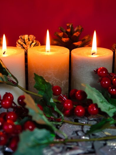 Three flaming candles stand between pine cones and red berries. The background is red and conveys a cozy, festive atmosphere. | © pixabay.com