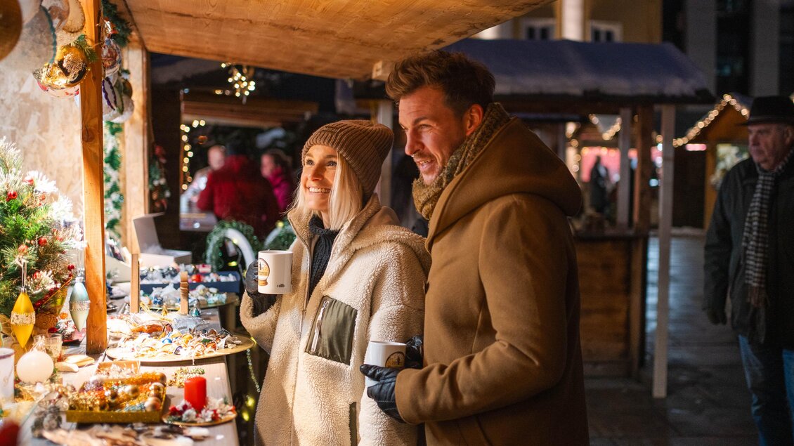Ein festlicher Weihnachtsmarkt mit leuchtenden Ständen und fröhlichen Menschen. Ein Paar genießt heiße Getränke und betrachtet die angebotenen Waren. | © Ekatarina Paller