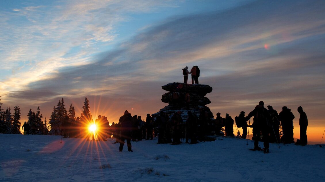 Eine Gruppe von Menschen steht im Schnee und beobachtet den Sonnenuntergang. Im Hintergrund sind Bäume und ein Hügel zu sehen. | © Kurt Elmleitner