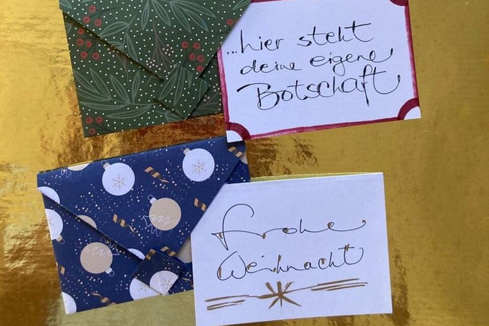 Drei dekorative Umschläge auf einem goldenen Hintergrund. Einer der Umschläge trägt die Aufschrift „Frohe Weihnachten“. | © AndersOrt Haus der Frauen