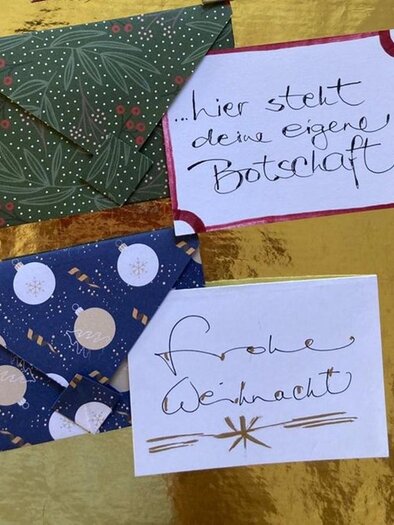 Drei dekorative Umschläge auf einem goldenen Hintergrund. Einer der Umschläge trägt die Aufschrift „Frohe Weihnachten“. | © AndersOrt Haus der Frauen