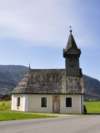 A charming chapel with a pointed steeple, surrounded by green meadows and mountains in the background. The sky is clear and blue. | © Steinegger