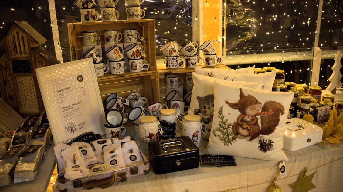 Ein festlich dekorierter Marktstand mit verschiedenen Tassen, Kissen und Marmeladengläsern. Im Hintergrund funkeln Lichter, die eine gemütliche Atmosphäre schaffen. | © Flourl´s Schenke