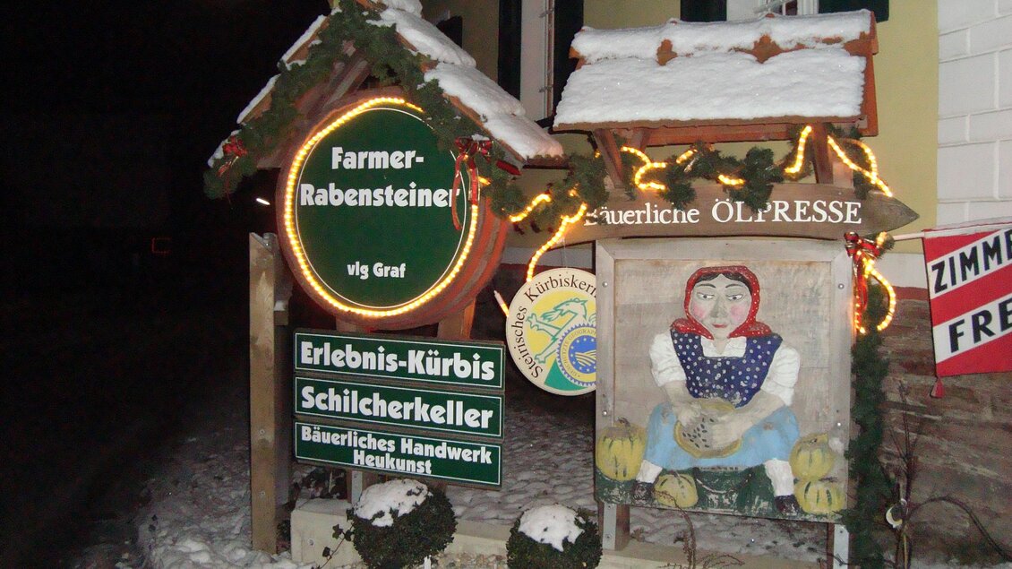 Ein festlich dekoriertes Schild mit Lichtern und einer Malerei zeigt eine typische Figur. Im Hintergrund liegt Schnee, und es deutet auf einen örtlichen Bauernladen hin.
