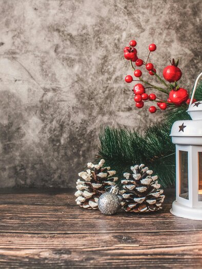 Eine weiße Laterne steht auf einem Holztisch, umgeben von Tannenzweigen, Tannenzapfen und roten Beeren. Die Laterne strahlt warmes Licht aus, das eine gemütliche Atmosphäre schafft. | © tijana-drndarski-unsplash