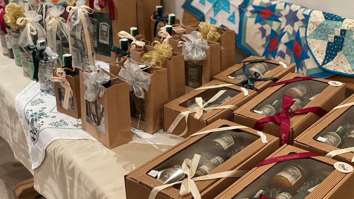 Ein Tisch mit verschiedenen schön verpackten Flaschen in Geschenkboxen. Die Flaschen sind mit Bändern und Stoff dekoriert, und im Hintergrund ist eine bunte Quilts sichtbar. | © Hofladen Hauptmann