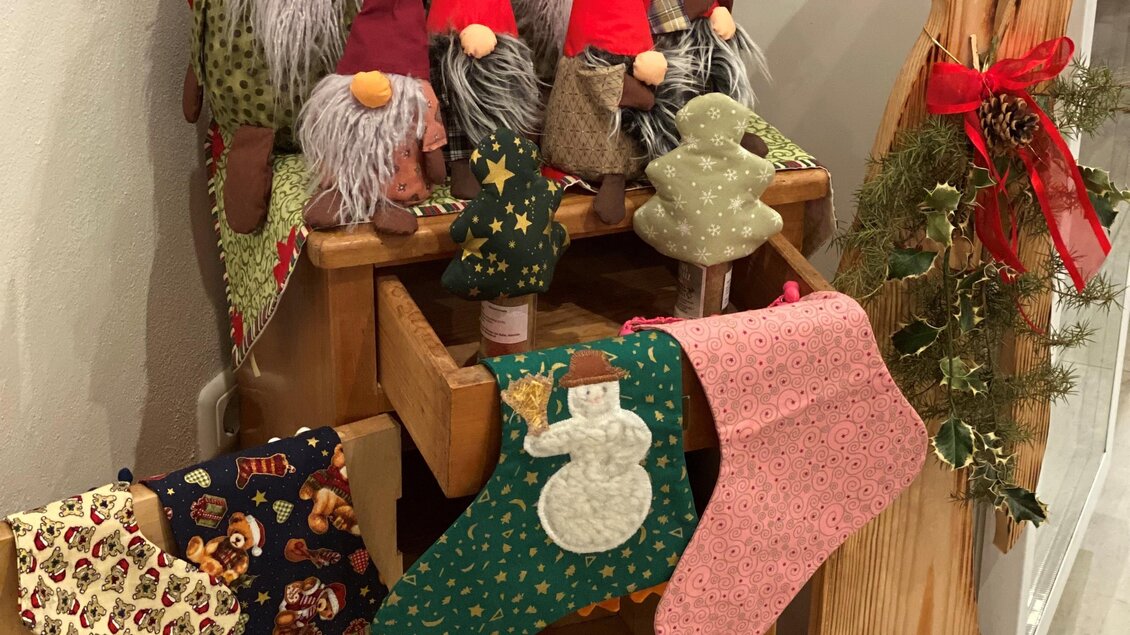 Eine festliche Weihnachtsdekoration mit mehreren Stoffgnomen und bunten Strümpfen. Im Hintergrund sind dekorative Elemente wie ein Kranz und ein Holzbrett zu sehen. | © Hofladen Hauptmann