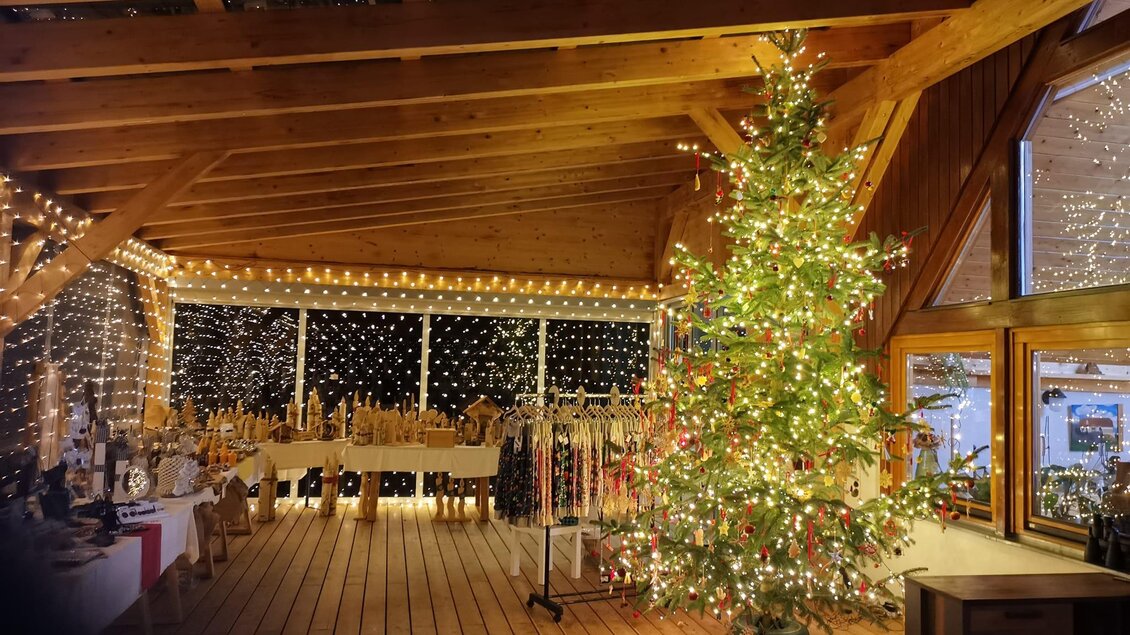 Ein gemütlicher Innenraum mit einem festlich geschmückten Weihnachtsbaum. Die Wände sind mit Lichtern dekoriert und es gibt Holzhandwerk auf den Tischen. | © Flourl´s Schenke