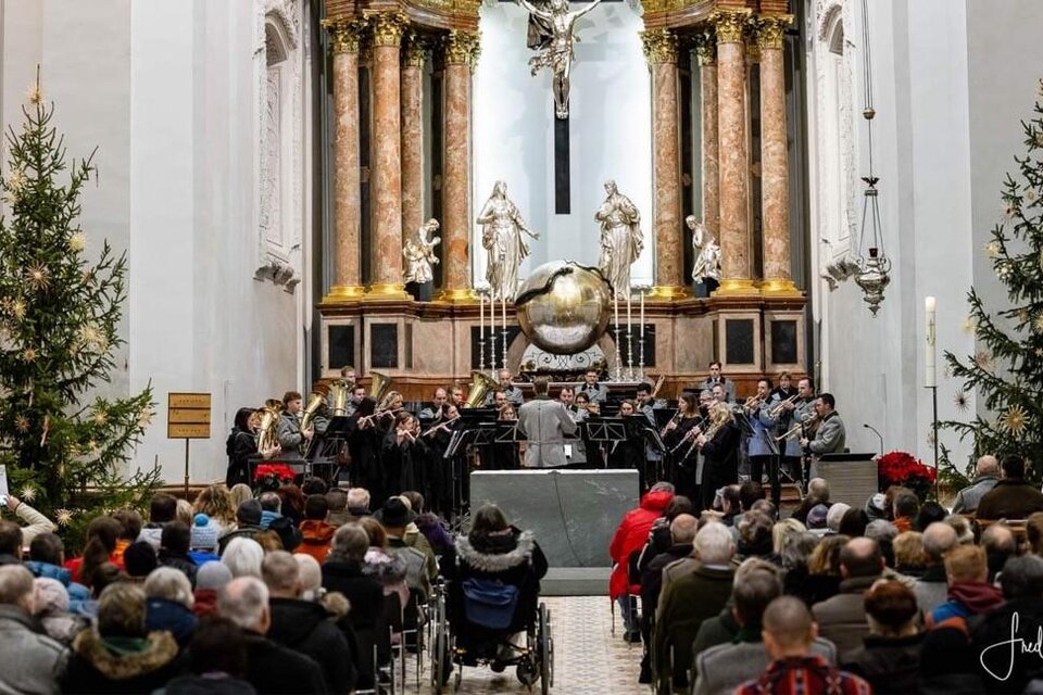 Ein Musikensemble spielt in einer Kirche vor einer großen Menschenmenge. Der Raum ist festlich geschmückt mit Weihnachtsbäumen. | © Stadtkapelle Mariazell, Fred Lindmoser