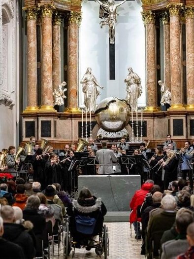 Ein Musikensemble spielt in einer Kirche vor einer großen Menschenmenge. Der Raum ist festlich geschmückt mit Weihnachtsbäumen. | © Stadtkapelle Mariazell, Fred Lindmoser