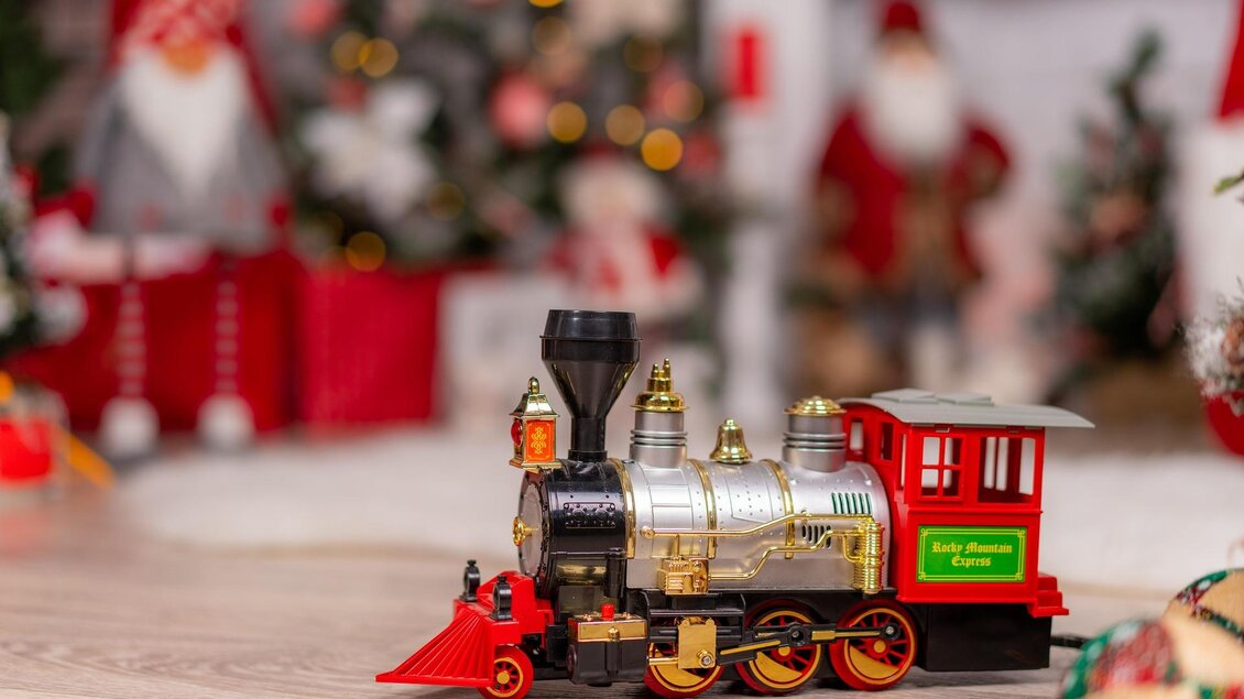 Eine glänzende Spielzeuglokomotive steht auf einem Holztisch. Im Hintergrund sind festliche Weihnachtsdekorationen und -figuren zu sehen. | © pixabay.com