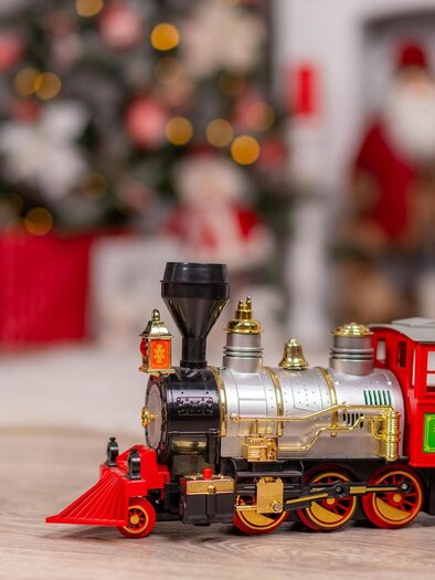 A shiny toy locomotive sits on a wooden table. In the background, festive Christmas decorations and figures can be seen. | © pixabay.com