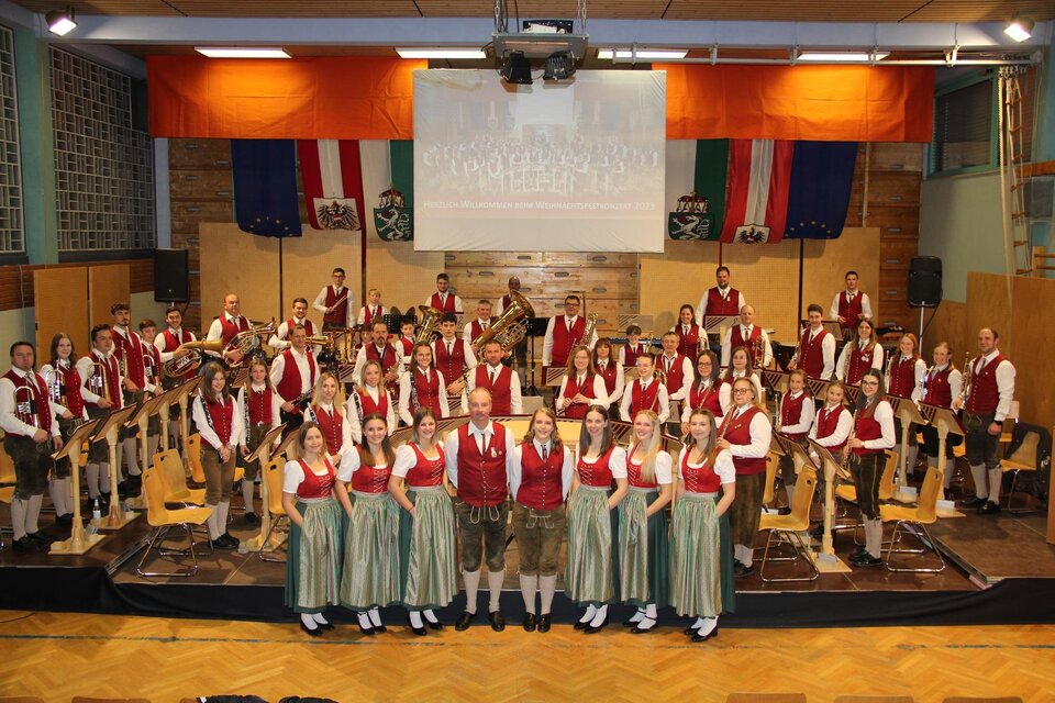 Eine große musizierende Gruppe in traditionellen österreichischen Trachten steht auf der Bühne. Im Hintergrund hängen Fahnen und ein Banner. | © Musikverein St. Lambrecht