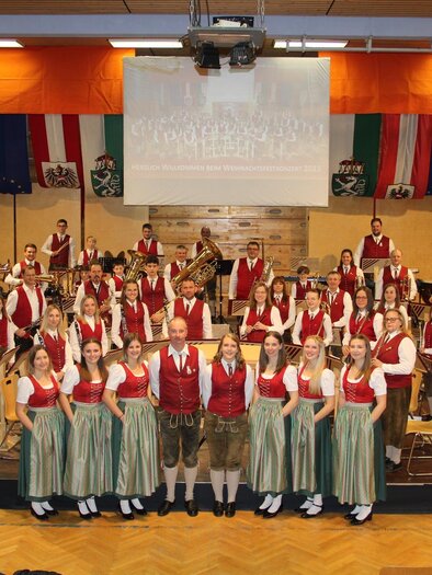 Eine große musizierende Gruppe in traditionellen österreichischen Trachten steht auf der Bühne. Im Hintergrund hängen Fahnen und ein Banner. | © Musikverein St. Lambrecht
