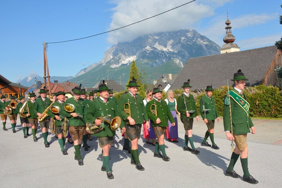 Eine Gruppe von Menschen in traditionellen Trachten marschiert eine Straße entlang. Im Hintergrund sind Berge und schöne Häuser sichtbar. | © Marktmusikkapelle Irdning