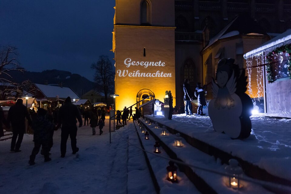 Eine festlich beleuchtete Winterlandschaft mit einem Kirchengebäude im Hintergrund. Menschen genießen den Weihnachtsmarkt bei Schneefall und sehen die Schrift "Gesegnete Weihnachten".