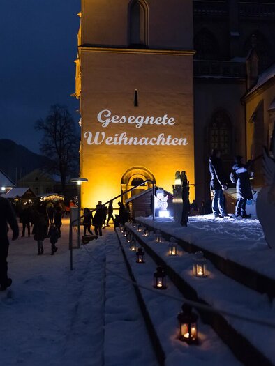 Eine festlich beleuchtete Winterlandschaft mit einem Kirchengebäude im Hintergrund. Menschen genießen den Weihnachtsmarkt bei Schneefall und sehen die Schrift "Gesegnete Weihnachten".