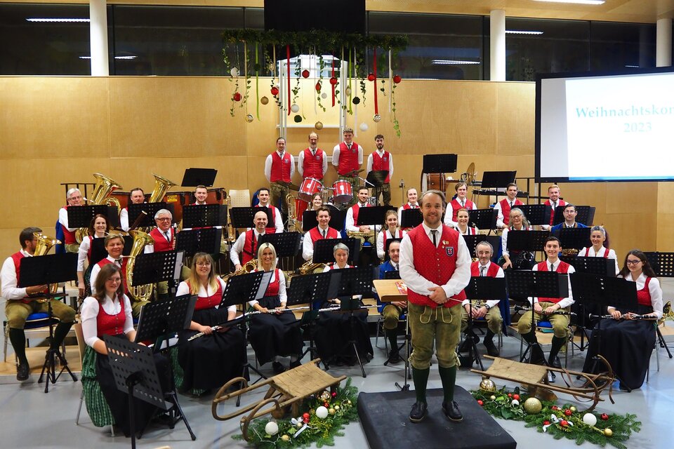 Eine große Blasmusikgruppe in traditionellen Trachten steht auf einer Bühne. Im Hintergrund hängen Weihnachtsdekorationen, und es sind Notenstände sichtbar. | © MV Weinitzen