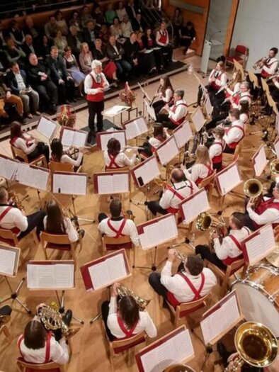 A large orchestra is playing on a stage. In the background, an audience is seated and listening attentively. | © Greith Haus