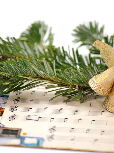 A small wooden angel sits on a sheet of music with Christmas carols. Below, fir branches lie, conveying a festive atmosphere. | © pixabay.com